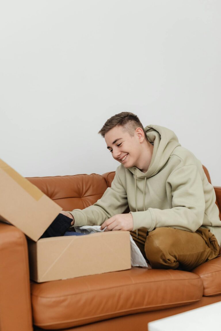 A teenager happily unboxes a package while sitting on a brown leather couch indoors.
