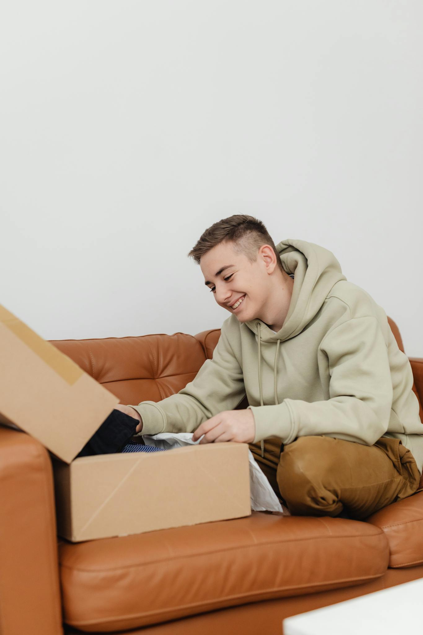 A teenager happily unboxes a package while sitting on a brown leather couch indoors.